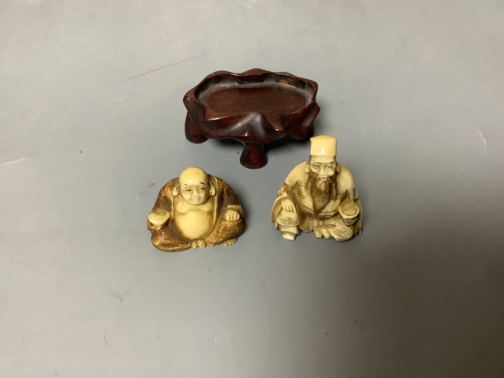 Two Japanese ivory netsuke of Hotei and a sage, Meiji period, wood stand